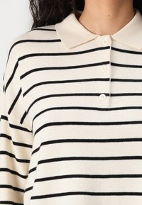 Striped pullover with a cream base and black horizontal stripes, featuring a collar and four buttons at the neckline. Soft texture.