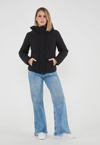 Giacca nera imbottita con cappuccio, dotata di tasche laterali e chiusura con zip. Indossata sopra jeans a gamba larga blu chiaro e sneakers bianche.