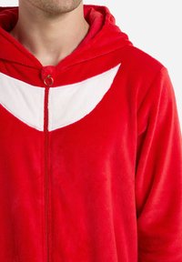 Red fleece onesie with hood, featuring a white accent on the chest, front zipper, and soft texture. Detailed stitching visible.