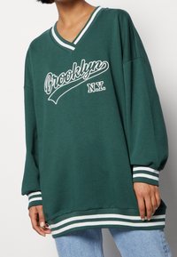 Sweat-shirt vert oversize avec impression blanche « Brooklyn N.Y. », encolure en V, poignets et ourlet rayés. Matière en coton, coupe décontractée.
