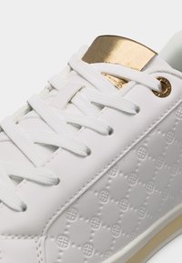 Λευκό αθλητικό παπούτσι με ανάγλυφο κυκλικό μοτίβο, λευκά κορδόνια και μεταλλικές χρυσές λεπτομέρειες στη γλωττίδα και τις τρύπες των κορδονιών.