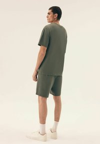 Krótkie rękawy oliwkowej zielonej dwuczęściowej odzieży wykonanej z miękkiego materiału. Luźny t-shirt w komplecie z dopasowanymi szortami; noszone z białymi trampkami i skarpetkami.
