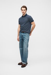 Μπλε ναυτικής ρίγας πόλο πουκάμισο συνδυασμένο με ανοιχτό μπλε τζιν παντελόνι κανονικής γραμμής. Το μοντέλο στέκεται με τα χέρια στις τσέπες, φορώντας καφέ παπούτσια. Καθαρό φόντο.