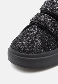 Punta di sneaker nera con superficie texturizzata glitterata e due ampie fasce in velcro su uno sfondo bianco.