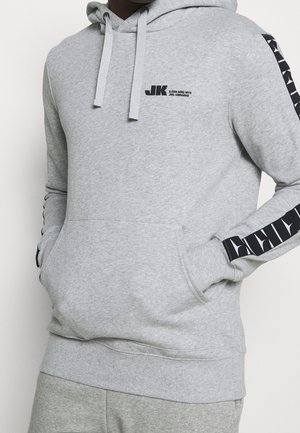 Person trägt hellgrauen Hoodie mit Fronttasche und schwarzem Logo-Text "JK Björn Borg mit Joel Kinnaman" auf der Brust und gemusterten schwarzen Streifen auf den Ärmeln.