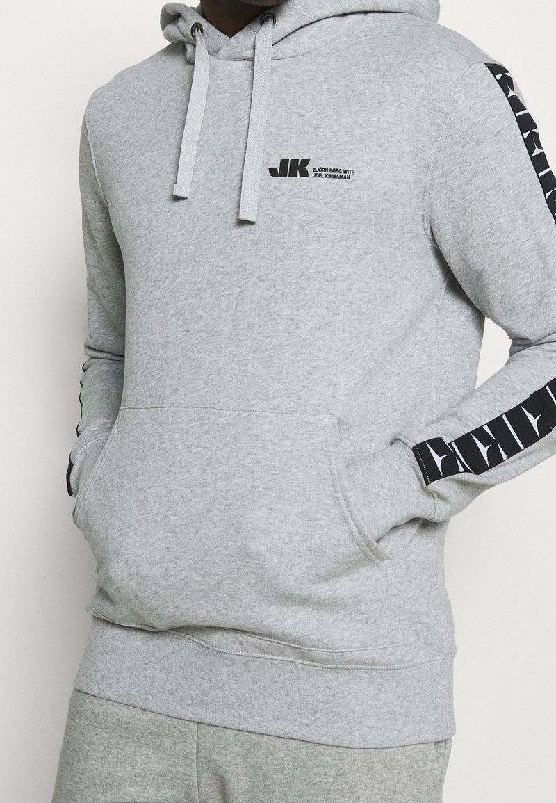 Personne portant un sweat à capuche gris clair avec une poche avant et le texte du logo noir "JK Björn Borg avec Joel Kinnaman" sur la poitrine ainsi que des rayures noires à motifs sur les manches.