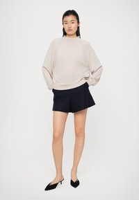 Maglione oversize beige chiaro con collo alto e maniche raglan, abbinato a pantaloni corti neri su misura e scarpe nere a punta.