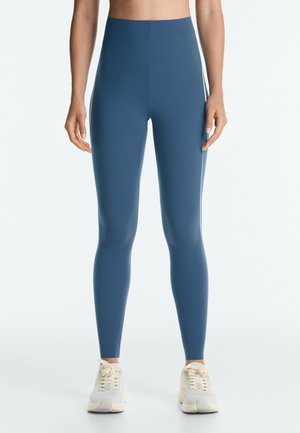 Persoon draagt blauwe legging met hoge taille en witte sneakers, staand met ontspannen armen langs het lichaam tegen een effen witte achtergrond.