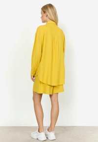 Gelbes Leinenhemd mit lockerer Passform, langen Ärmeln und abgerundetem Saum, kombiniert mit passenden Shorts. Weiße Sneaker runden das Outfit ab.