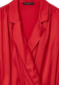 Blusa rossa di raso con scollo colletto, caratterizzata da dettagli plissettati sulle spalle e una superficie liscia e lucida. Etichetta di taglia S visibile.