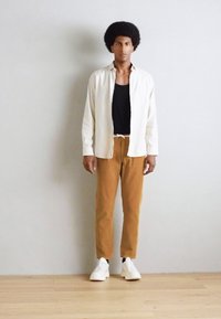 Witte button-up shirt over een zwarte tanktop, gecombineerd met een paarse corduroy broek en witte sneakers. Eenvoudige, casual outfit tegen een neutrale achtergrond.