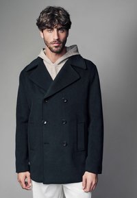 Dunkelgrauer Wollpea coat mit doppelreihiger Front, großen Revers und zwei vorderen Taschen. Über einem hellgrauen Hoodie getragen. Insgesamt elegantes Design.