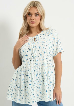 Femme aux longs cheveux blonds portant un chemisier blanc à manches courtes avec un motif floral bleu et un détail de nouage à l'avant, assorti à un jean bleu.