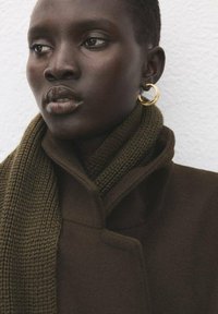 Brązowy płaszcz z wysokim kołnierzem, teksturowany oliwkowy szalik i złote kolczyki koła. Cera gładka, makijaż o neutralnych tonach.