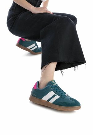 Baskets teal avec des rayures blanches et des accents rose, dotées d'une semelle en caoutchouc текстurée et de lacets. Portées avec un pantalon en denim noir raccourci.