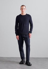 Pull en tricot noir avec ourlet côtelé, associé à un pantalon sombre. Le mannequin se tient devant un fond neutre, portant des chaussures noires.
