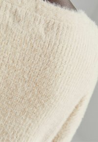 Svetřík krémové barvy s měkkým, strukturovaným pleteným vzorem, širokým výstřihem a viditelnými stehy podél švů.