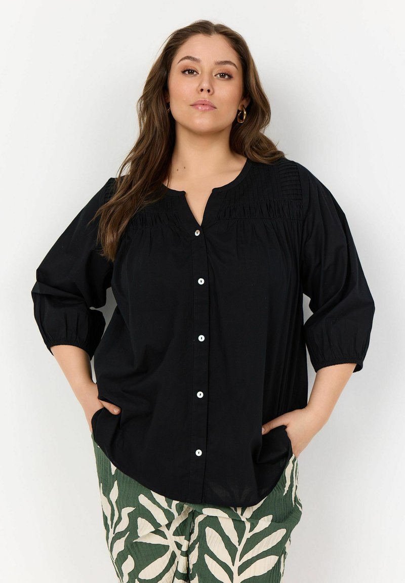 Camicia nera con colletto a V, maniche a tre quarti, dettagli arricciati sulle spalle e bottoni bianchi. Abbinata a pantaloni verdi con motivo.
