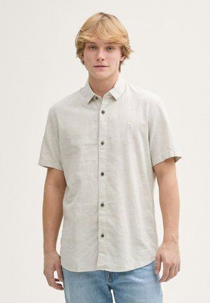 Camisa corta de manga en gris claro con un diseño de botones, que presenta botones oscuros, un cuello y un sutil logo en el lado izquierdo del pecho. Combinada con unos vaqueros de mezclilla azules.
