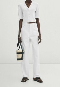 Weißes, geripptes, kurz geschnittenes Hemd mit Knöpfen, kombiniert mit hoch taillierten weißen Hosen. Schwarze flache Sandalen und eine kleine, gewebte Tasche mit schwarzen Akzenten.