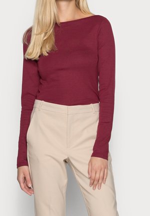 Kvinde iført en langærmet bordeaux trøje og beige bukser, stående med hænderne afslappet ved siden.