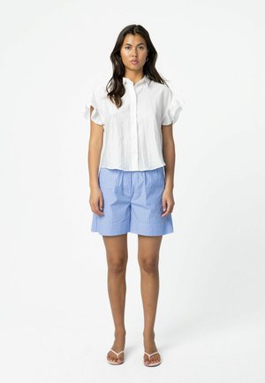 Vrouw met lang donker haar, gekleed in een wit kort mouwen overhemd met knopen, blauw-witte gestreepte korte broek en witte sandalen, staand en recht vooruit kijkend.