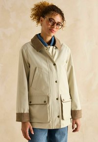 Jachetă impermeabilă bej cu guler din catifea, dotată cu nasturi tip snap, două buzunare frontale și o croială lejeră. Detalii din țesătură texturată.
