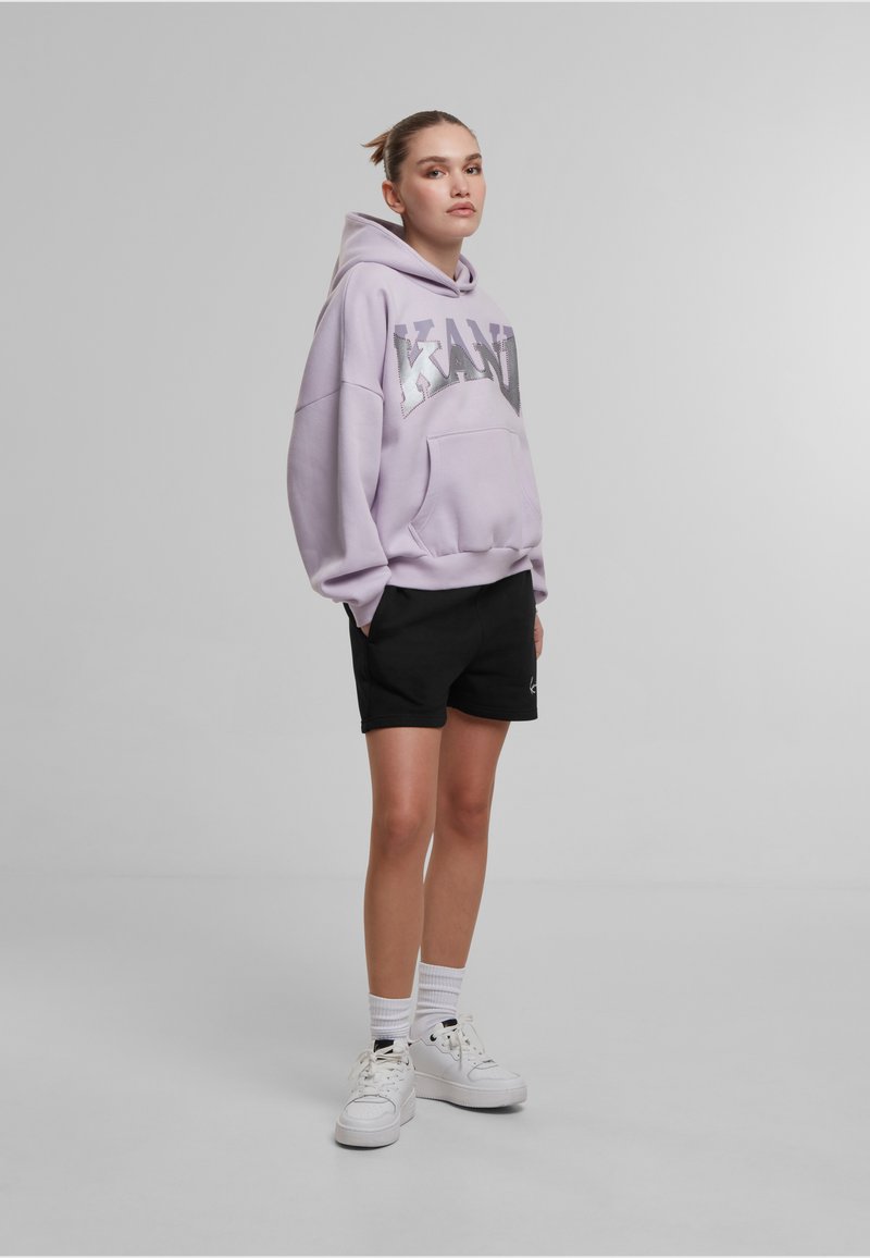 Lila oversized dukserica s kapuljačom s metalik natpisom "KANU", uparena s crnim šortsevima i bijelim tenisicama. Model stoji pred neutralnom pozadinom.