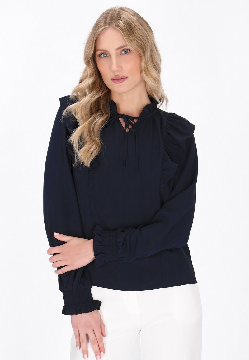 Blusa blu navy con dettagli arricciati, scollatura con fiocco e polsini arr gathered. Tessuto morbido con una finitura liscia, adatta per un abbigliamento casual o formale.