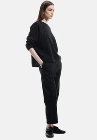 Czarny oversize sweter z teksturowanym wykończeniem, zestawiony z czarnymi zwężającymi się spodniami. Model nosi czarne trampki, ręce w kieszeniach, widok z boku.