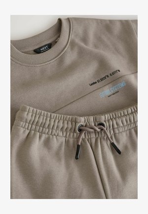 Bēša kokvilnas džemperis un atbilstošas bikses. Džemperim ir ribota apkakle, drukāts teksts un logo. Biksēm ir elastīga jostasvieta ar auklu.