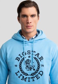 Svetle modrá mikina s kapucí vyrobená z bavlny, s černou grafikou lva a textem "BIGSTAR LIMITED" v kruhovém designu. Šňůrky na kapuci.