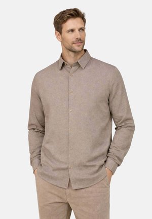 Uomo con capelli castani corti che indossa una camicia beige a maniche lunghe con bottoni e pantaloni abbinati, con le mani in tasca, guarda verso destra.