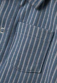 Tessuto a strisce blu e beige con finitura testurizzata, dotato di tasca sul petto e chiusura con bottone, con dettagli di cucitura visibili.