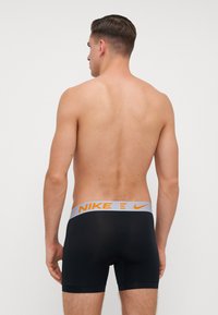 Melnas bokseršortus ar pelēku Nike jostasvietu, kurā ir oranži uzraksti, skatīts no aizmugures uz balta fona.