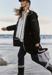 Giacca nera impermeabile con fodera bianca, abbinata a jeans scuri e stivali in gomma neri con strisce bianche. Include una borsa grigia.