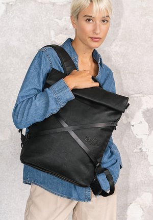Young woman with short blonde hair wearing a blue denim jacket carries a large black leather backpack with roll-top closure and cross straps.