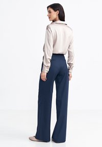 Beżowa blouse o jedwabnym wykończeniu z kołnierzykiem na plecach; granatowe spodnie szerokie w paski. Gładka tekstura, luźny krój, mankiety zapinane na guziki oraz wysoki stan.