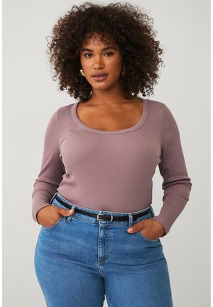 Kvinde med krøllet hår iført en mauve langærmet top stoppet ned i blå jeans med et sort bælte, stående med hænderne i lommerne.