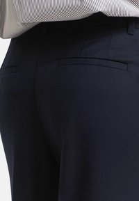 Nahaufnahme von dunkelblauen maßgeschneiderten Hosen mit aufgesetzten Gesäßtaschen, kombiniert mit einem in die Hose gesteckten hellgestreiften Hemd.
