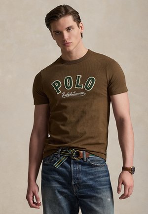 Ung mand iført en brun Polo Ralph Lauren t-shirt, blå jeans, stribet bælte og et armbåndsur, stående mod en neutral baggrund.