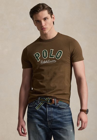 Νέος άνδρας που φοράει καφέ t-shirt Polo Ralph Lauren, μπλε τζιν, ριγέ ζώνη και ρολόι χειρός, στέκεται με φόντο ουδέτερο.