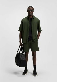 Olivgrön kortärmad skjorta och matchande shorts, med en svart t-shirt under. Svart väska och sneakers, med mörka solglasögon.