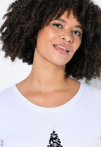 Hvid T-shirt med rund halsudskæring og et trædesign dekoreret med pailletter. Stoffet ser blødt ud med en glat tekstur.