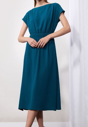 Femme portant une robe midi teal avec des manches courtes et une taille élastique, se tenant avec les mains jointes devant, sur fond blanc.