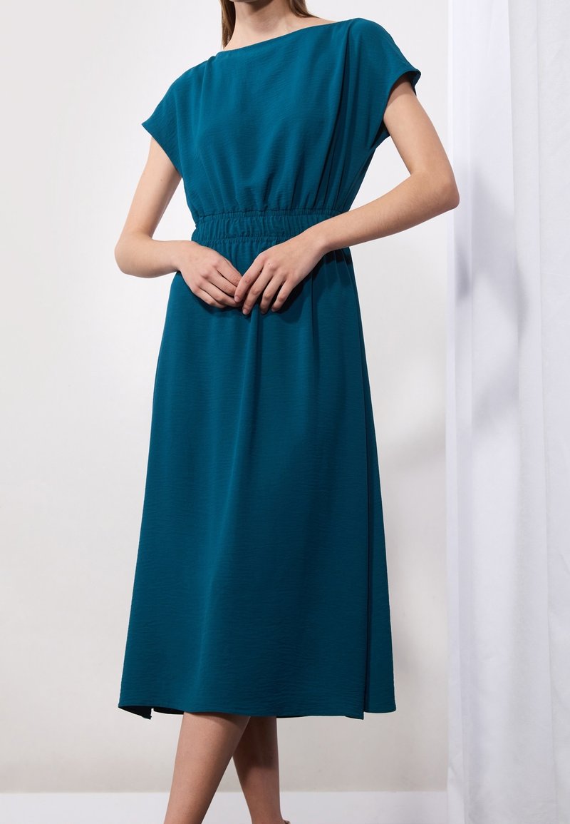 Femme portant une robe midi teal avec des manches courtes et une taille élastique, se tenant avec les mains jointes devant, sur fond blanc.