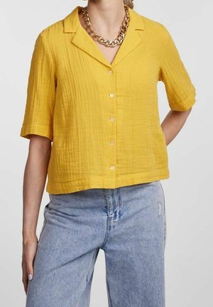 Frau trägt eine kurzärmelige, gelbe Bluse mit Knöpfen und Kragen, kombiniert mit hellblauen, hoch taillierten Jeans und einer goldenen Halskette.