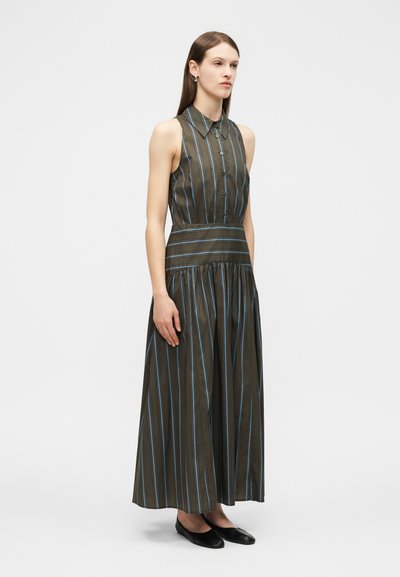 Γυναίκα που φοράει αμάνικο μάξι φόρεμα ελιάς με κάθετες λεπτές μπλε ρίγες, μπούστο με γιακά και κουμπιά, και μαύρα φλατ, στέκεται πλαγίως.