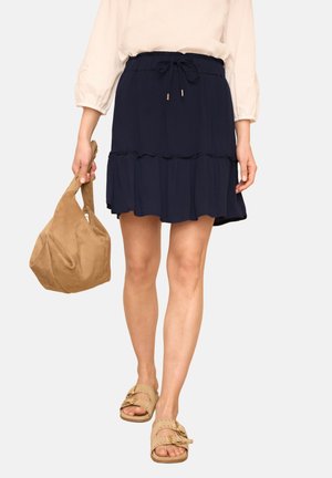 Kvinde iført en navyblå flæsekjol, holdende en tan ruskindstaske og iført beige sandaler med spænde, beskåret midt på torsoen.