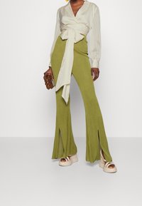 Blouse cache-cœur plissée crème à manches longues, associée à un pantalon évasé côtelé vert avec des fentes latérales. Le mannequin porte des sandales plateformes beige épaisses.